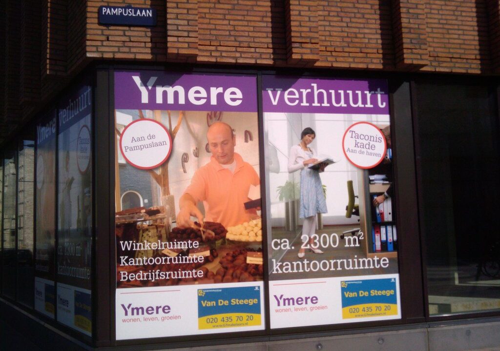 In de etalages van winkels hangen advertenties voor kantoor- en winkelruimtes te huur in Ymere. De linkerposter toont een man die brood schikt, de rechterposter een vrouw met papieren. Borden en contactgegevens zijn zichtbaar.
