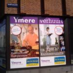 In de etalages van winkels hangen advertenties voor kantoor- en winkelruimtes te huur in Ymere. De linkerposter toont een man die brood schikt, de rechterposter een vrouw met papieren. Borden en contactgegevens zijn zichtbaar.