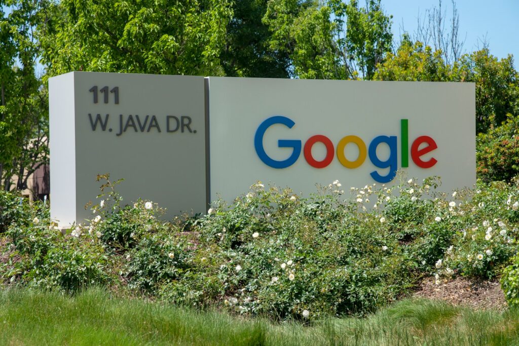 Op een wit buitenbord staat links de tekst "111 W. JAVA DR." en rechts het kleurrijke Google-logo, omringd door groene struiken, witte bloemen en bomen op de achtergrond.