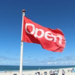 Een rode vlag met het woord "Open" in witte letters wappert aan een stok op een zonnig strand. Op de achtergrond zijn de oceaan, het zand en een aantal strandstoelen te zien, tegen een strakblauwe lucht.