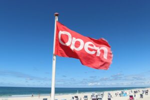 Een rode vlag met het woord "Open" in witte letters wappert aan een stok op een zonnig strand. Op de achtergrond zijn de oceaan, het zand en een aantal strandstoelen te zien, tegen een strakblauwe lucht.
